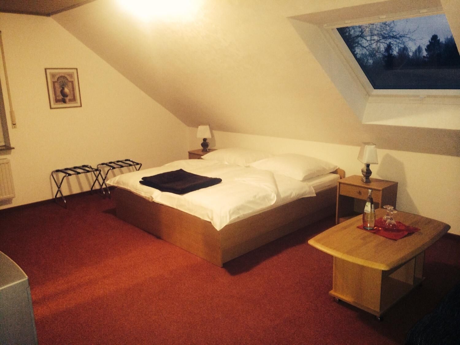Una cama individual en una habitación con techo inclinado y tragaluz. Hay una pequeña mesita de noche con una lámpara, un revistero de madera y un paño rojo sobre la mesa.