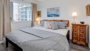 Et soveværelse med seng, et natbord, en kommode, et vindue med gardiner og et billede på væggen.