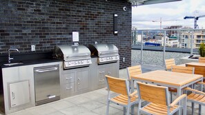 Une cuisine extérieure équipée de deux barbecues, d’un évier et d’un égouttoir, installée contre un mur de briques et offrant une vue sur une marina et des bâtiments en arrière-plan.