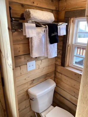 A wooden cabin bathroom with a toilet, a towel rack with several towels, and a window. - A View for Two: Tiny House with BIG VIEWS (Fleetwood)