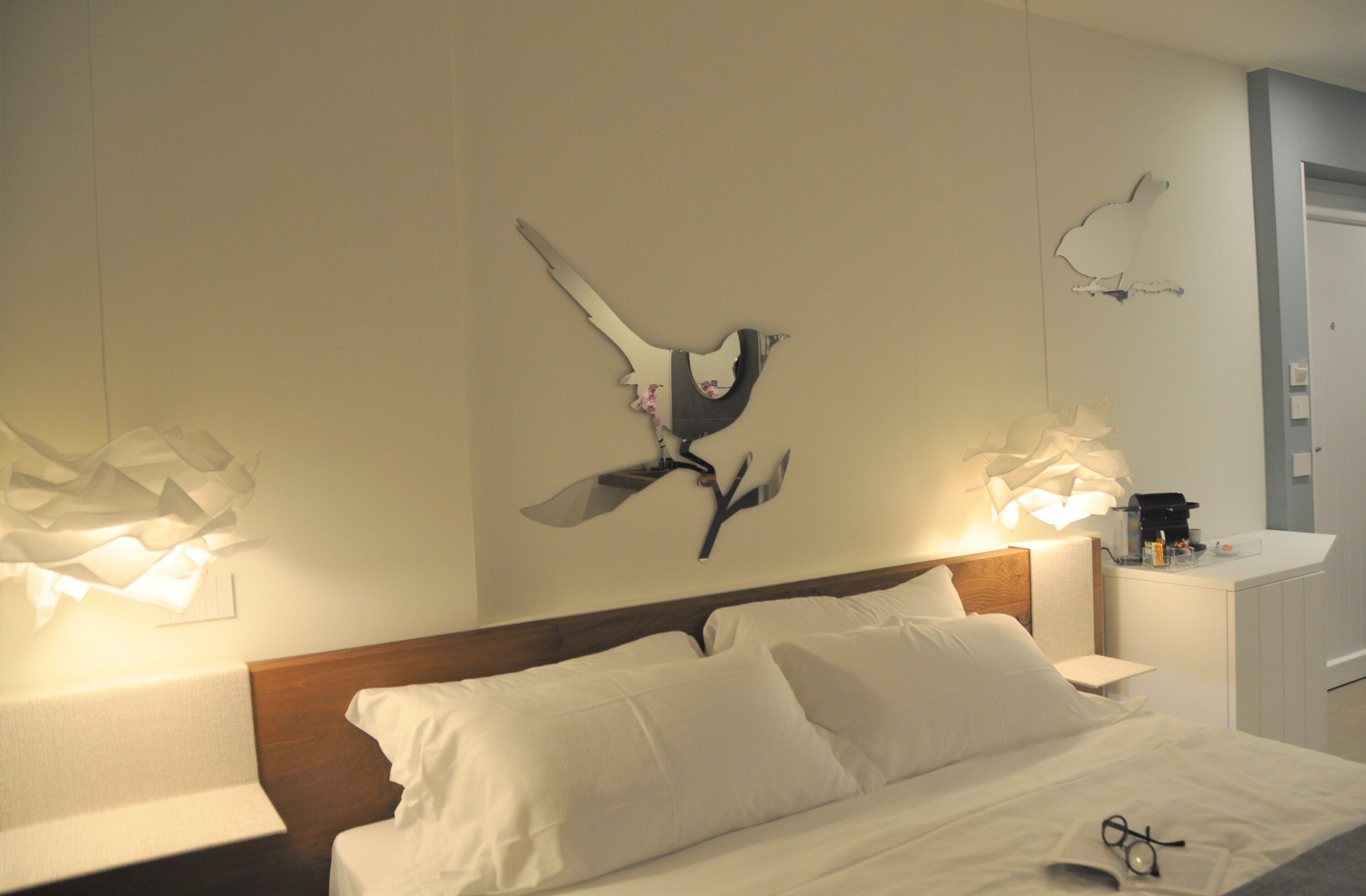A hotel room with a neatly made bed, a wall-mounted artwork of a bird, and a small bedside table with a lamp.