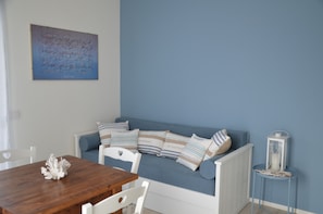 Quarto com sofá azul, cadeiras brancas, mesa de madeira e uma pintura abstrata pendurada na parede.