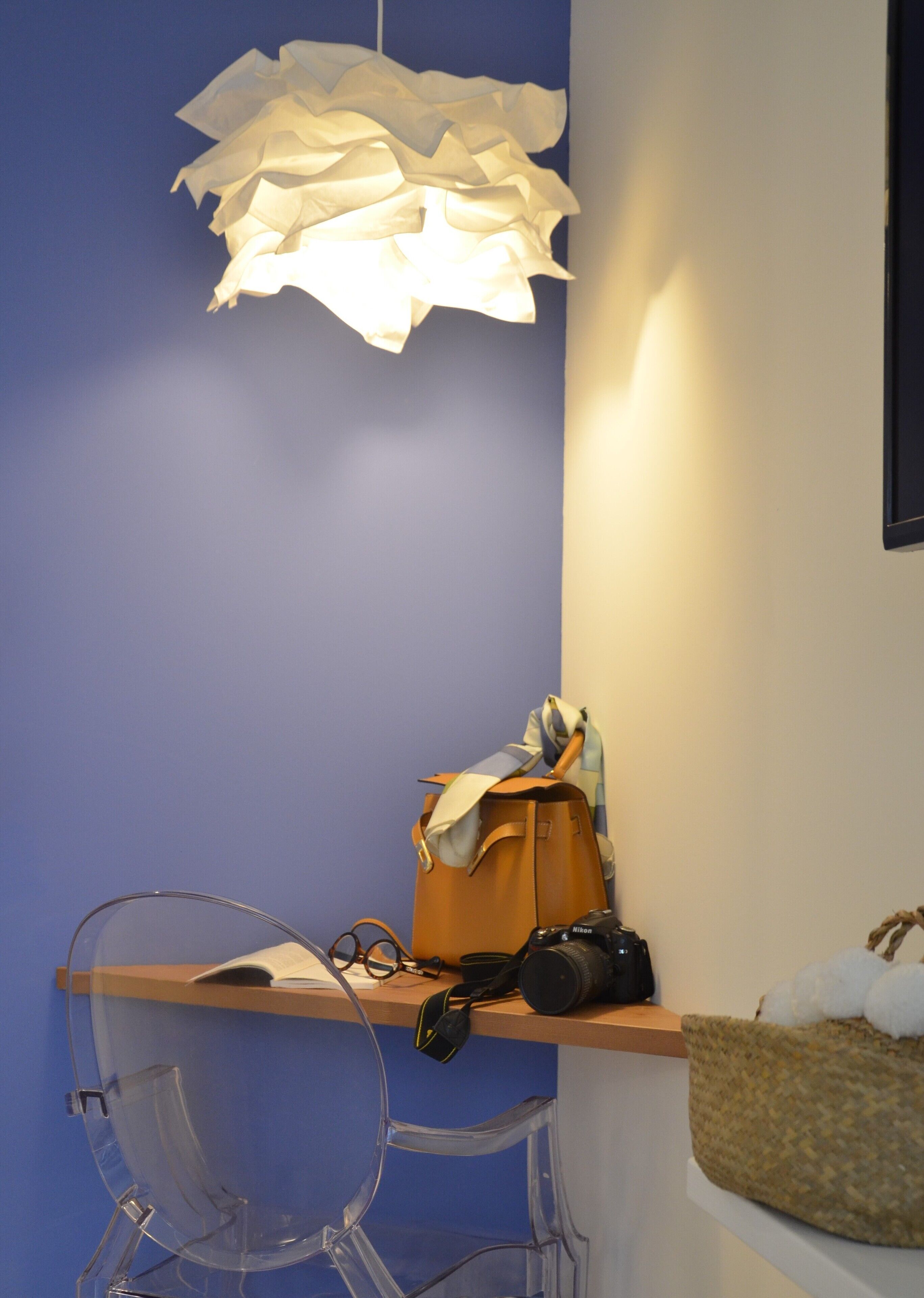Une pièce avec un mur bleu, une chaise en plastique transparent, un bureau en bois, un sac à main marron, un appareil photo Nikon et un panier en osier.