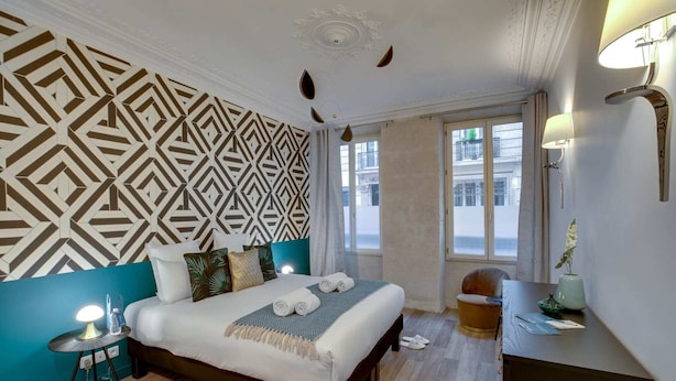 A hotel room with a geometric patterned wall, a bed with a white duvet and multiple pillows, a small bedside table with a lamp, a wooden floor, and a window with a view of buildings. - Sweett | Trevise - Three Bedroom Apartment, Sleeps 6 (Paris)