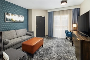 Een hotelkamer met een bank, poef, een flatscreen televisie, bureau en een raam met gordijnen.