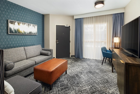 A hotel room with a sofa, ottoman, a flat-screen TV, a desk, and a window with curtains.
