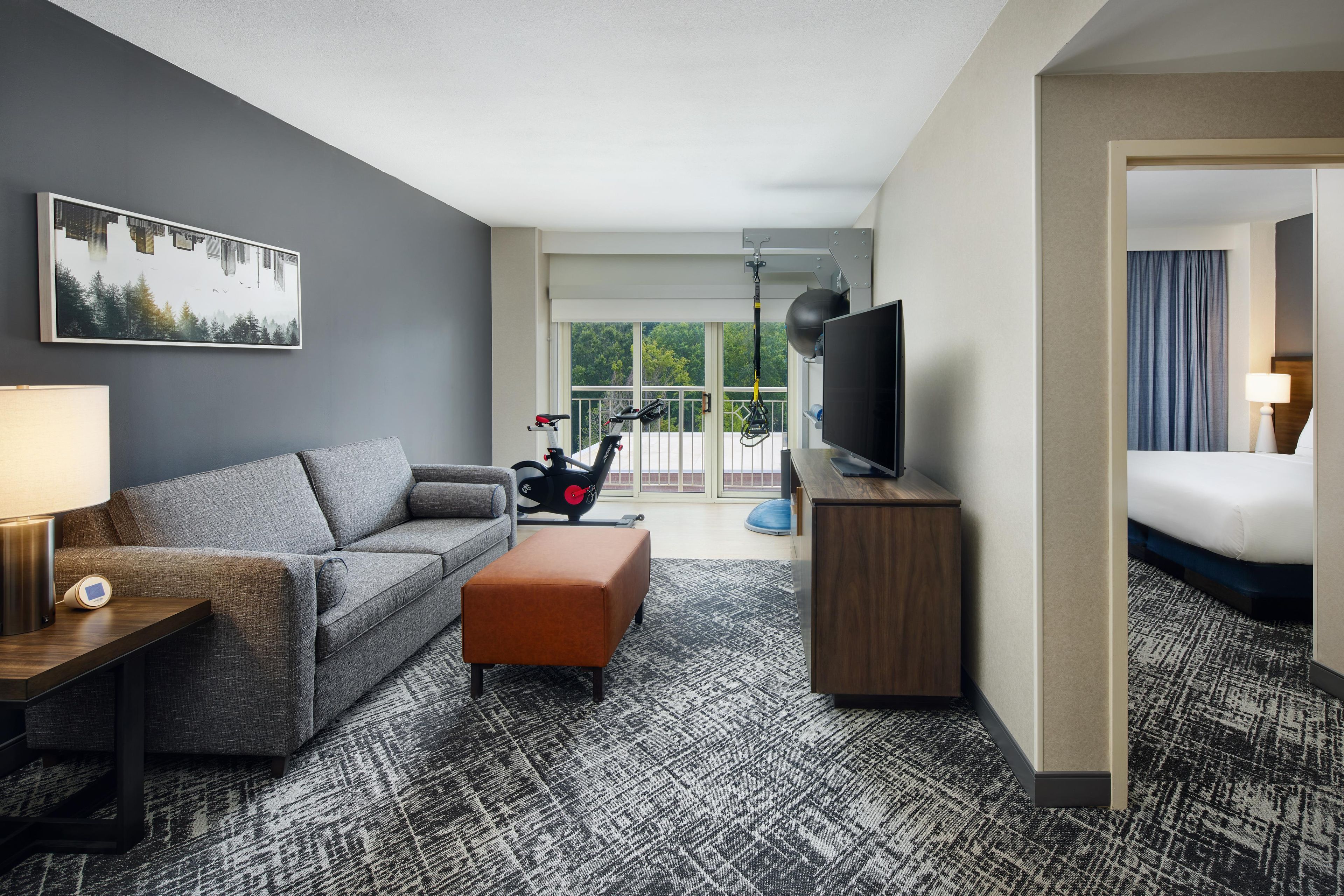 Habitación de hotel con sofá gris, un pequeño reposapiés, un televisor sobre un mueble de madera y un ventanal con vistas a árboles.