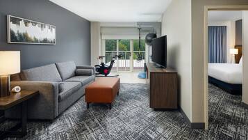 Habitación de hotel con sofá gris, un pequeño reposapiés, un televisor sobre un mueble de madera y un ventanal con vistas a árboles.