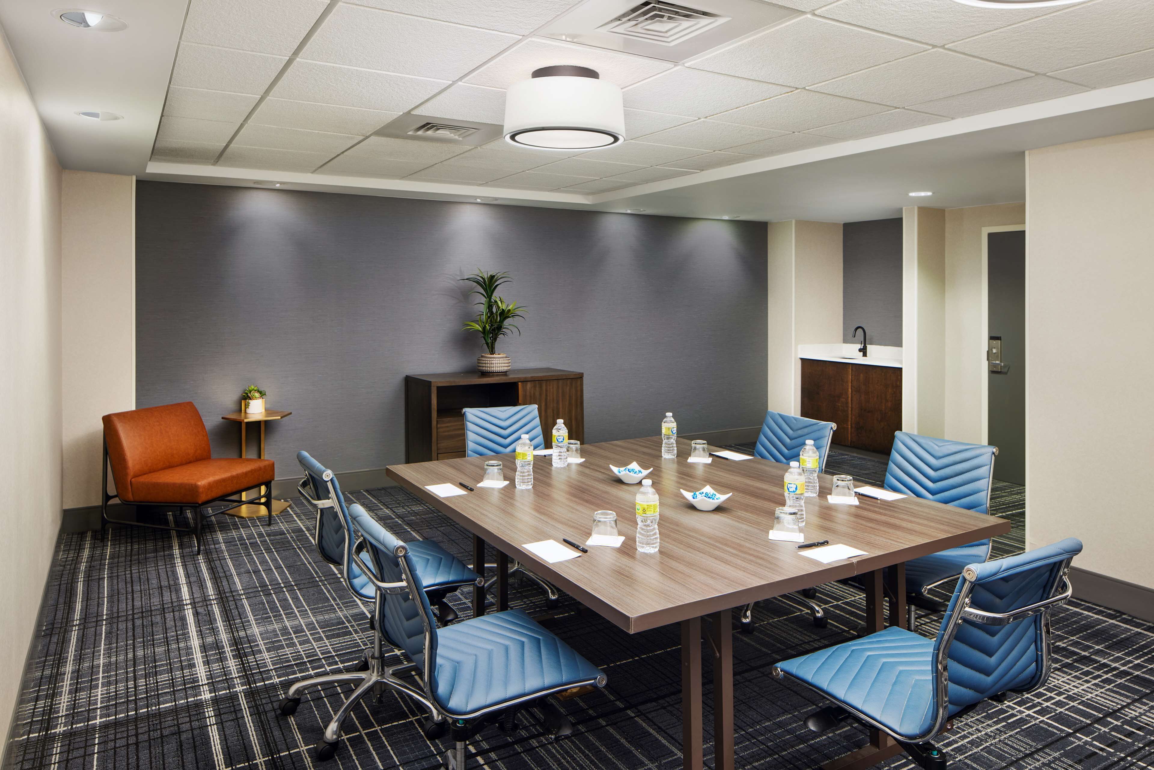 A conference room with a long table, blue chairs, a small orange chair, a potted plant, and a sink in the corner.
