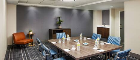 A conference room with a long table, blue chairs, a small orange chair, a potted plant, and a sink in the corner.