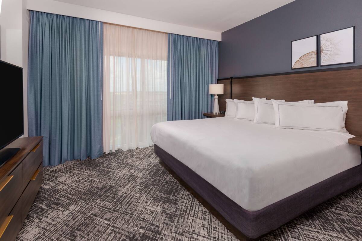 Une chambre d’hôtel avec un grand lit, un téléviseur à écran plat, une commode en bois et une fenêtre avec des rideaux bleus.
