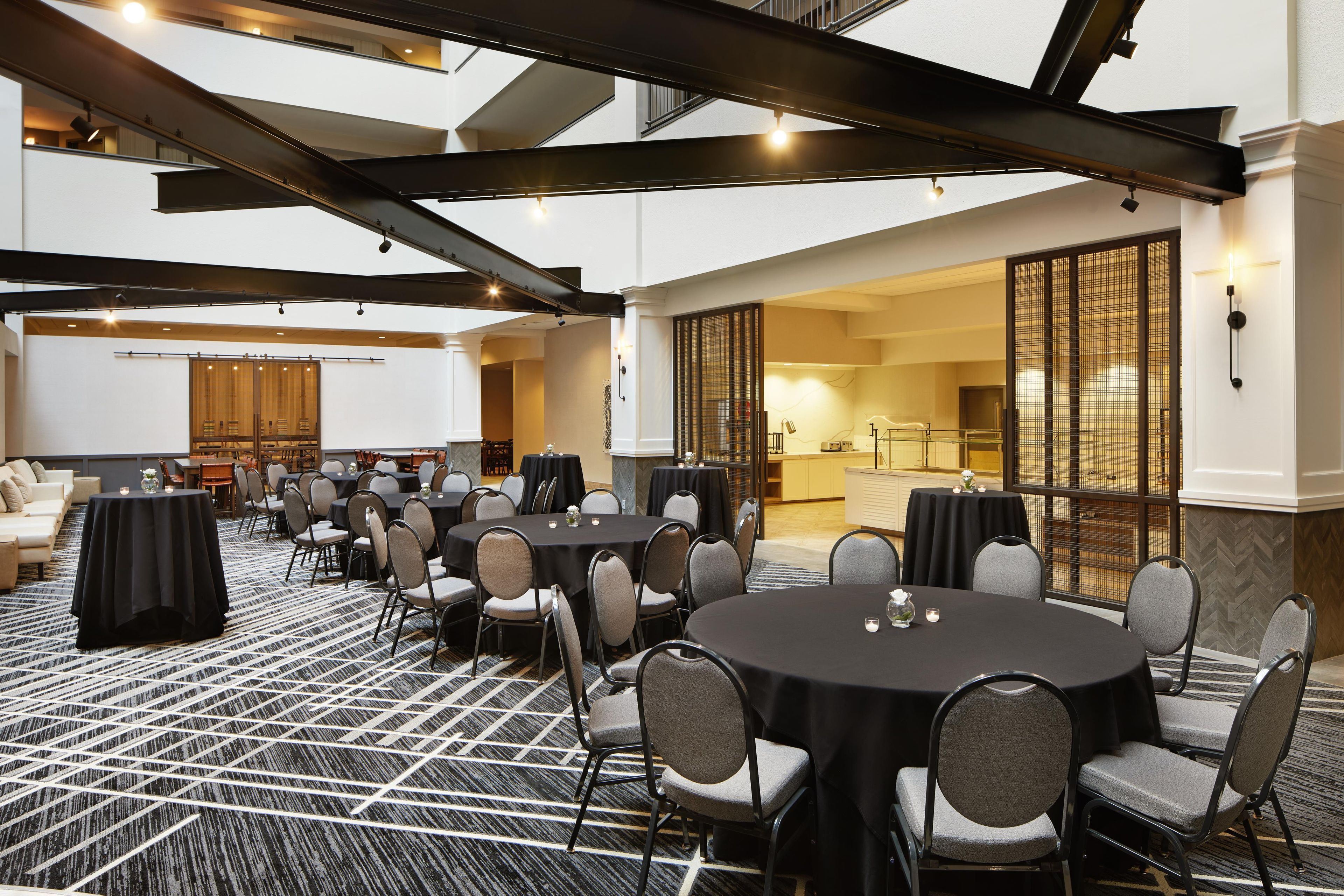 A conference room with round tables and chairs, a patterned carpet, and a view of a kitchen area.