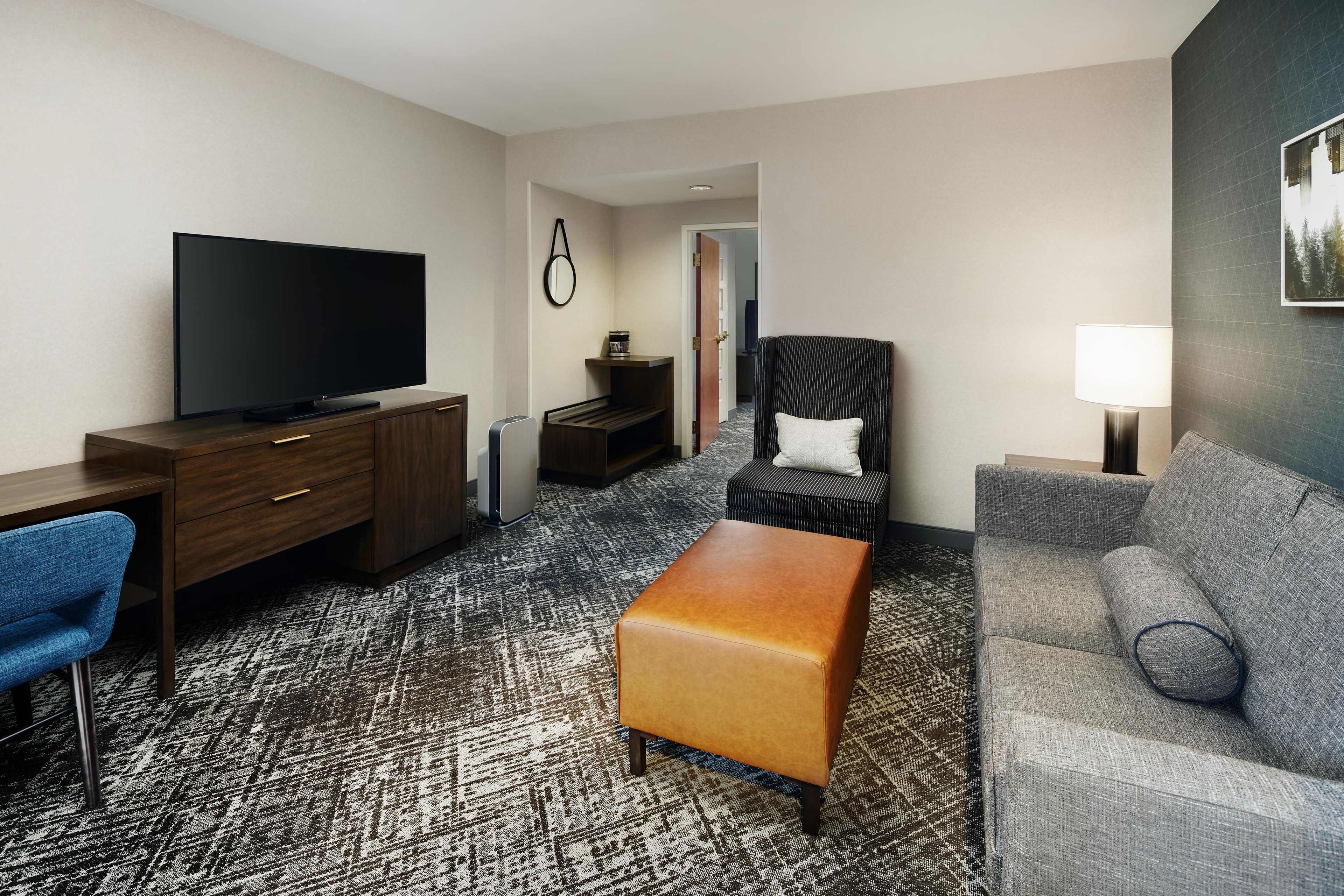 A hotel room with a flat-screen TV, a desk, a chair, a sofa, an ottoman, and a lamp.