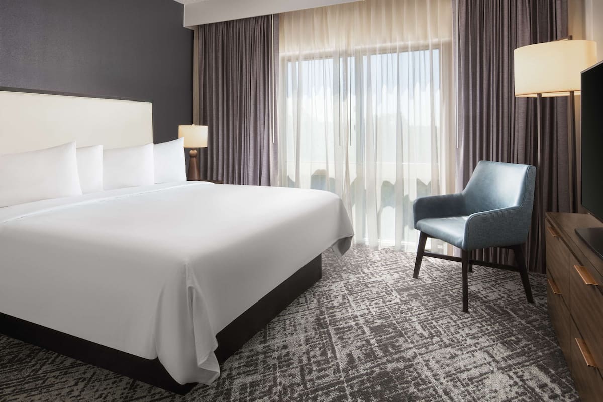 Une chambre d’hôtel avec un grand lit, une chaise, une télévision et une fenêtre avec des rideaux.