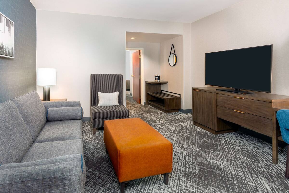 Une chambre d’hôtel avec un canapé gris, un pouf orange, un téléviseur à écran plat, un bureau et une chaise.