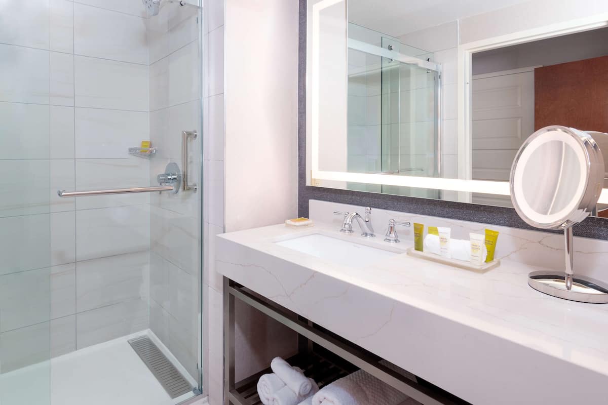 Une salle de bain moderne dotée d’une douche à parois de verre, d’un lavabo blanc avec un plan de toilette en marbre et d’un grand miroir à cadre rond.