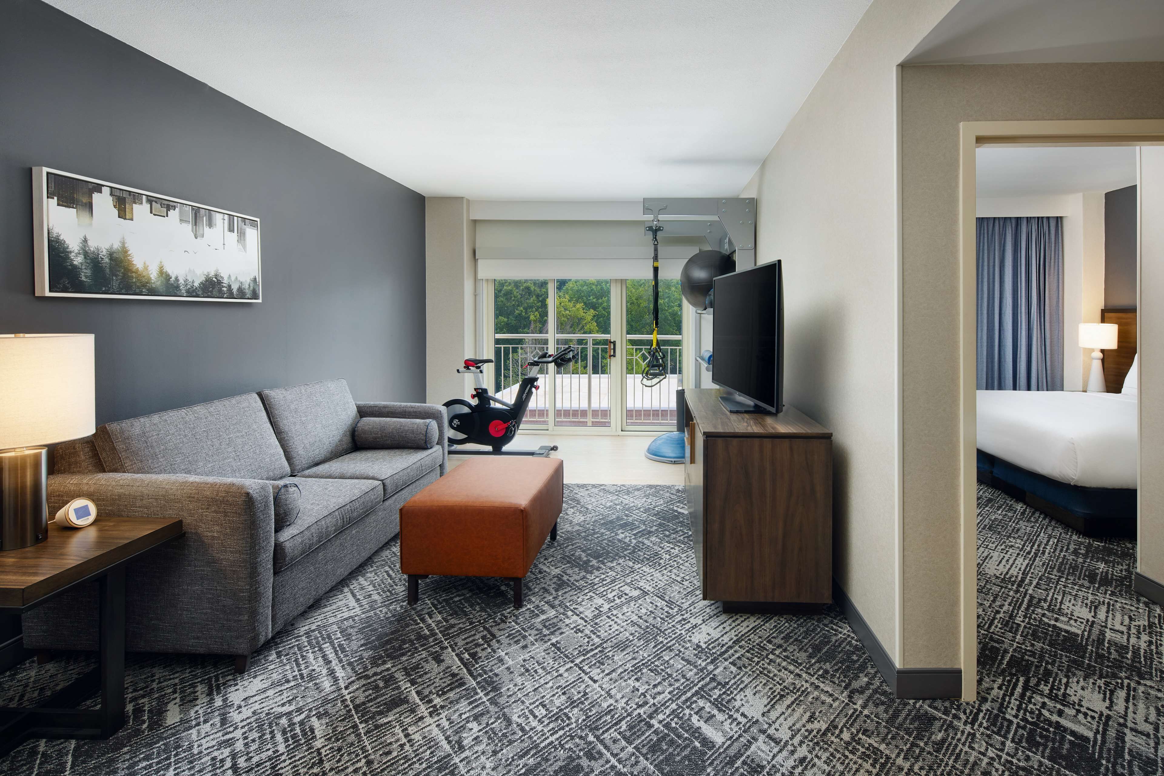 A hotel room with a grey sofa, a small orange ottoman, a TV on a wooden stand, a workout bike, and a balcony with a view of trees.