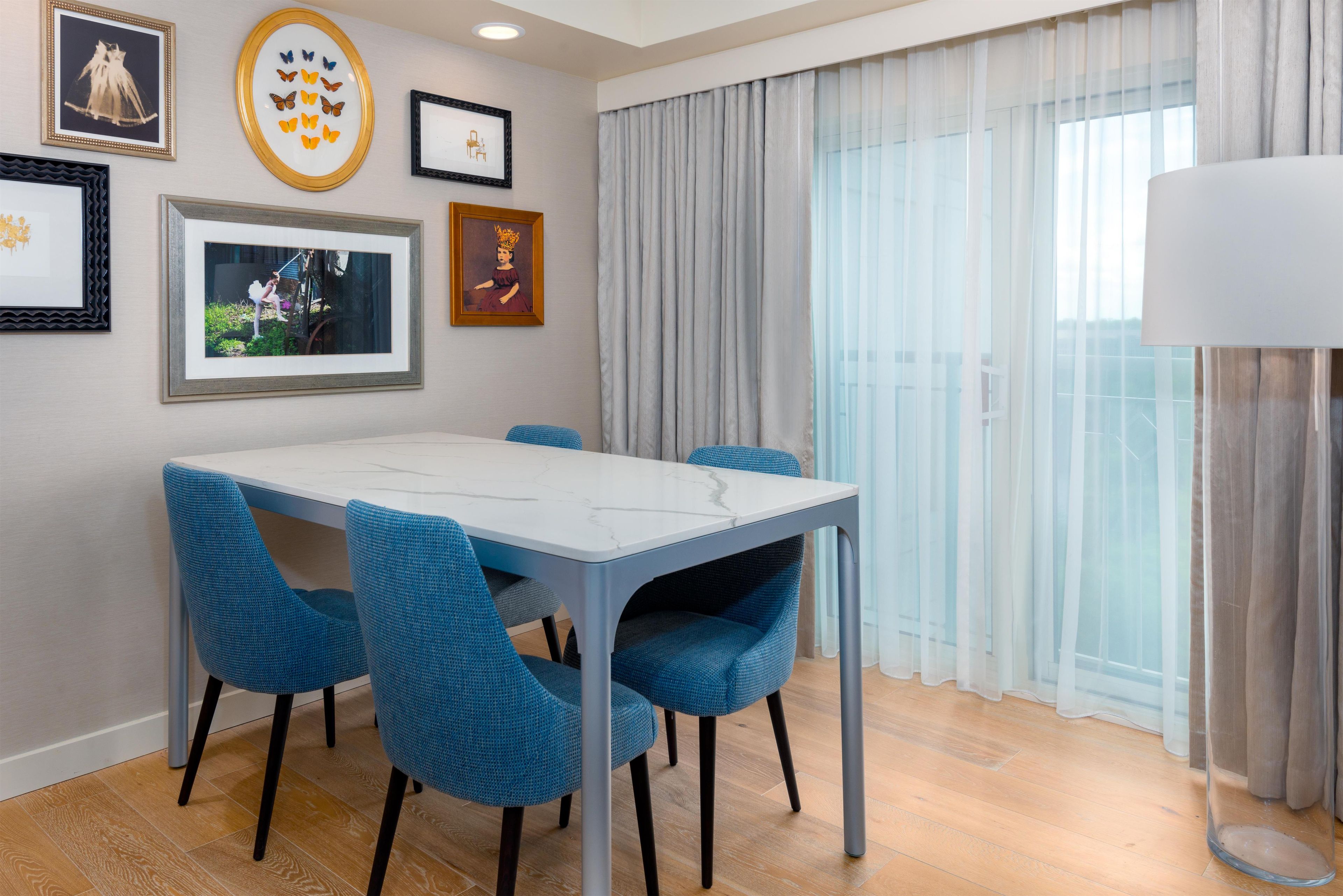 A dining area with a white table and blue chairs, framed artwork on the wall, and a large window with curtains.