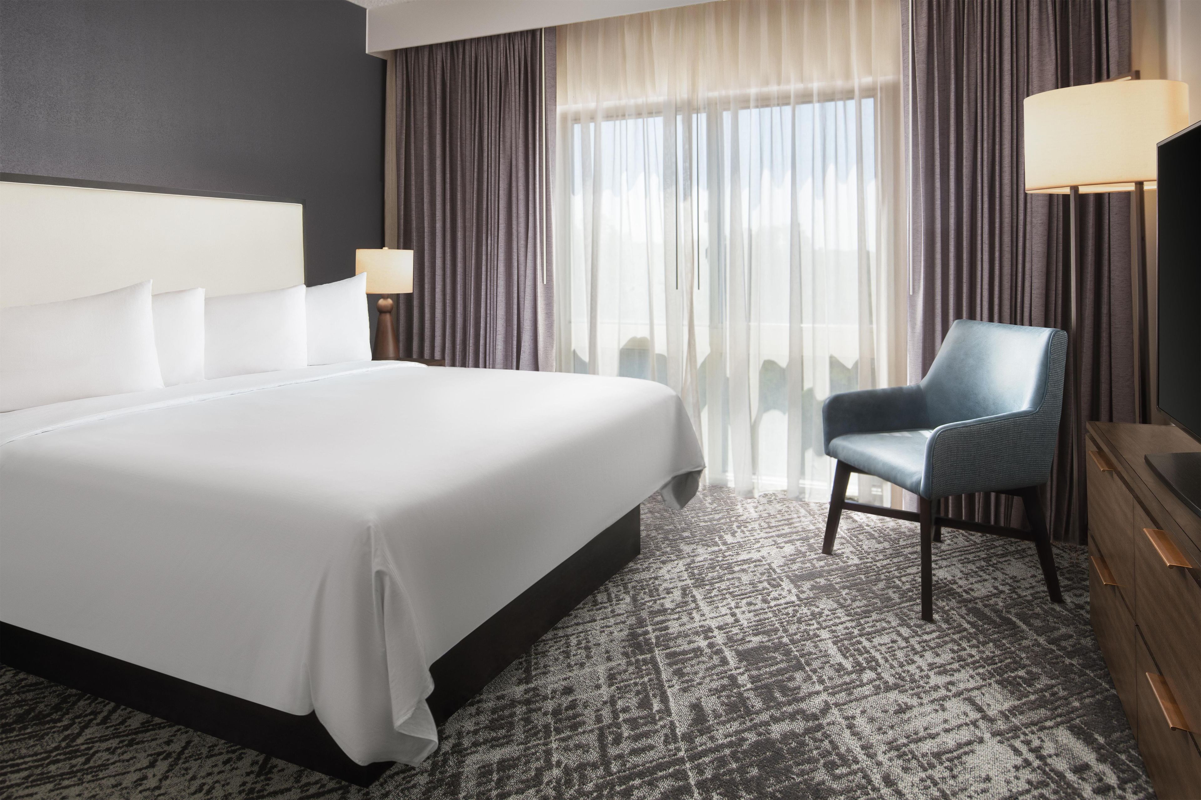 A hotel room with a large bed, a chair, a desk, and a view of the outdoors through sheer curtains.