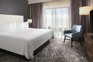 A hotel room with a large bed, a chair, a desk, and a view of the outdoors through sheer curtains.