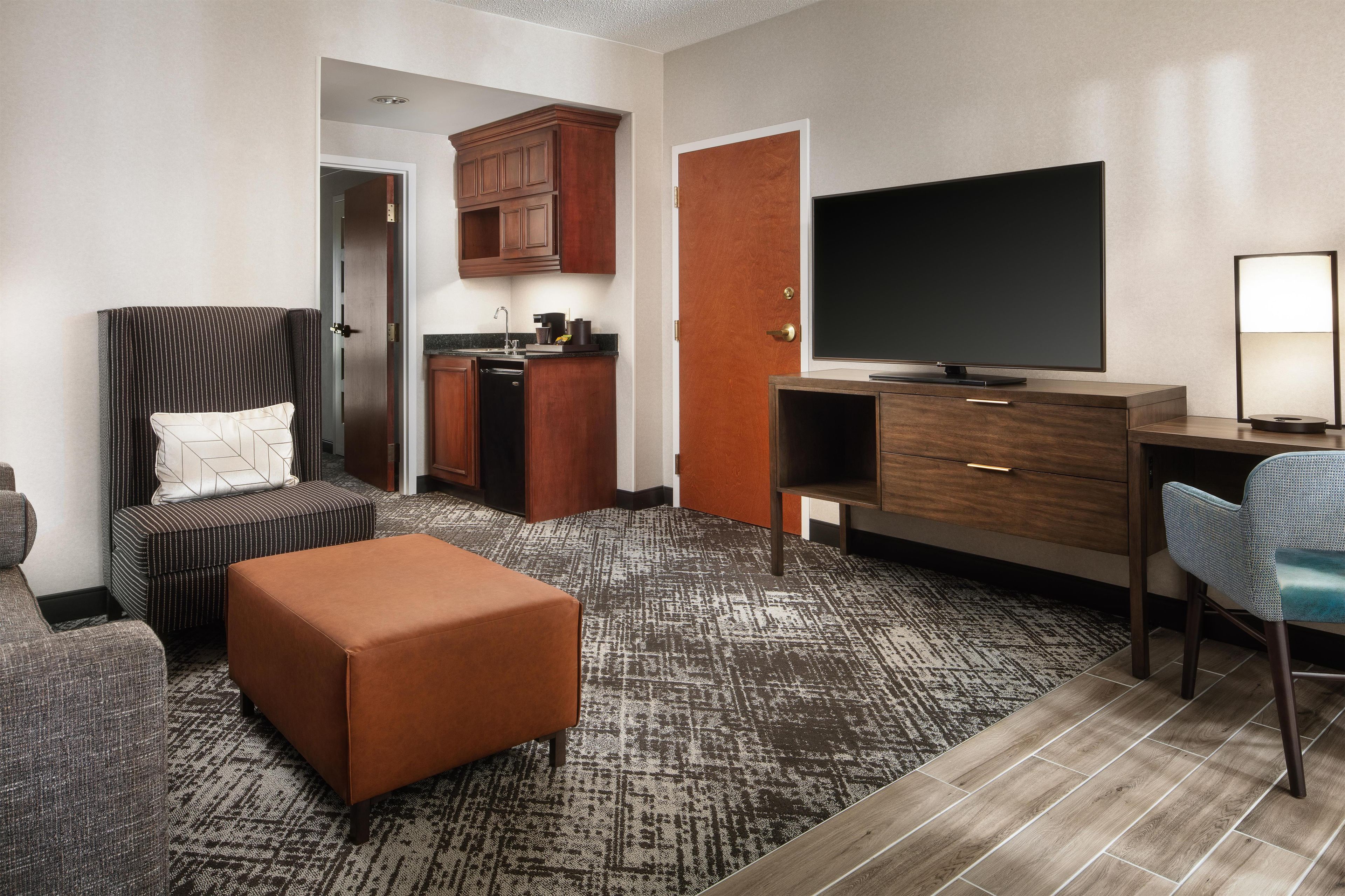 A hotel room with a sofa, ottoman, kitchenette, TV, desk, and a patterned carpet.