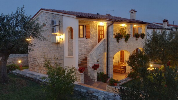 Una casa de piedra con una escalera que lleva a una puerta de madera, rodeada de follaje e iluminación exterior.