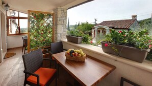 Un balcón con sillas de mimbre, una mesa de madera con una cesta de frutas, plantas en macetas y un muro de piedra.