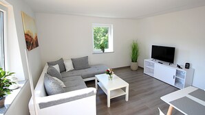 Un salón moderno con un sofá seccional gris, una mesa de centro blanca, un televisor de pantalla plana sobre un mueble blanco y una ventana con vistas a árboles.