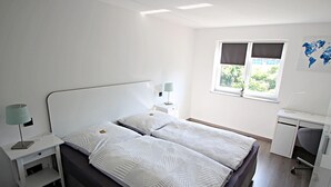 Un dormitorio ordenado con cama, mesitas de noche, escritorio y ventana con cortinas.