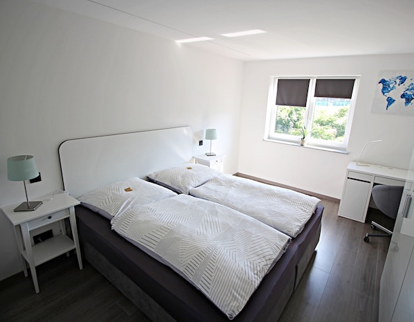 A neatly arranged bedroom with a bed, bedside tables, a desk, and a window with curtains. - Uraha Apartments (Herzogenaurach)