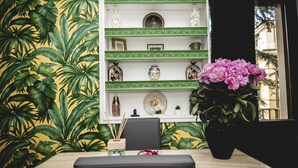 A room with a wooden table, a black chair, a green patterned shelf with decorative items, and a large potted plant with pink flowers.