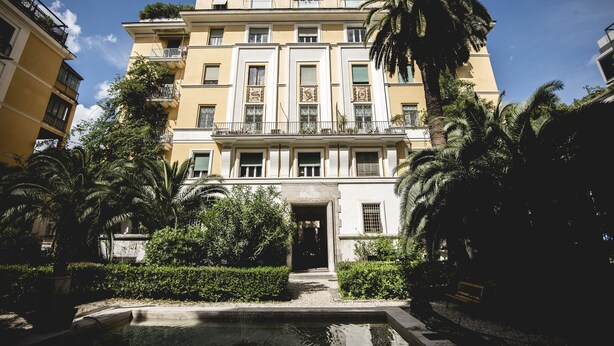 A multi-story building with balconies, a central entrance, and surrounding greenery.