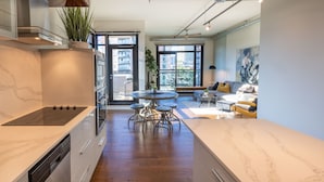 A modern kitchen with a built-in stove, a dining area with a round table and stools, and a living room with a sofa and a painting. - Jukebox Lofty Views-Perfect Mix of Indoor / Outdoor Living, Gym and Free Parking by Victoria Prime! (Victoria)