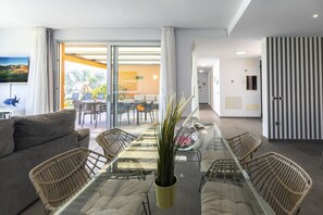 A modern living room with a glass dining table, wicker chairs, a sofa, and a large window with curtains.