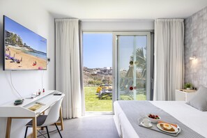 A hotel room with a bed, a desk, a chair, a TV, and a balcony with a view of the beach.