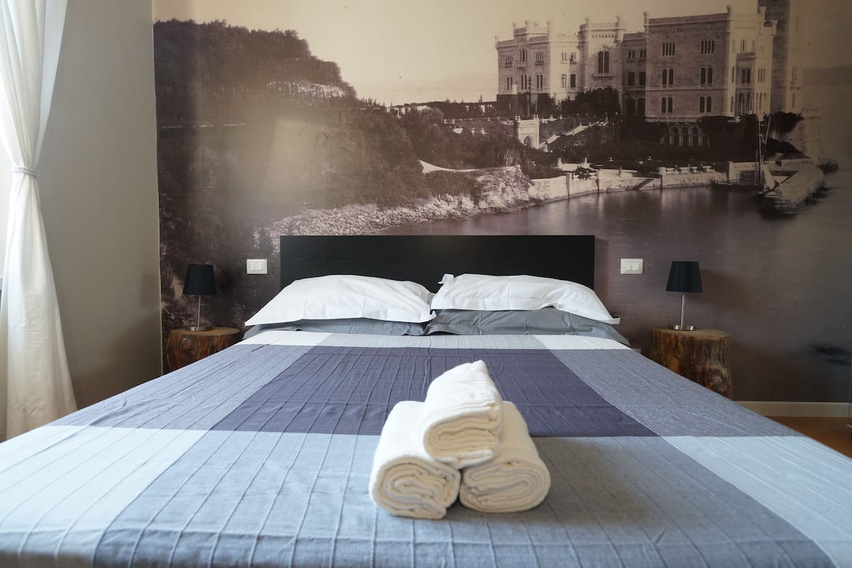Uma cama bem arrumada com colcha azul e branca listrada, duas almofadas brancas e toalhas enroladas sobre a cama. Há um mural grande na parede que retrata um edifício histórico e um rio.