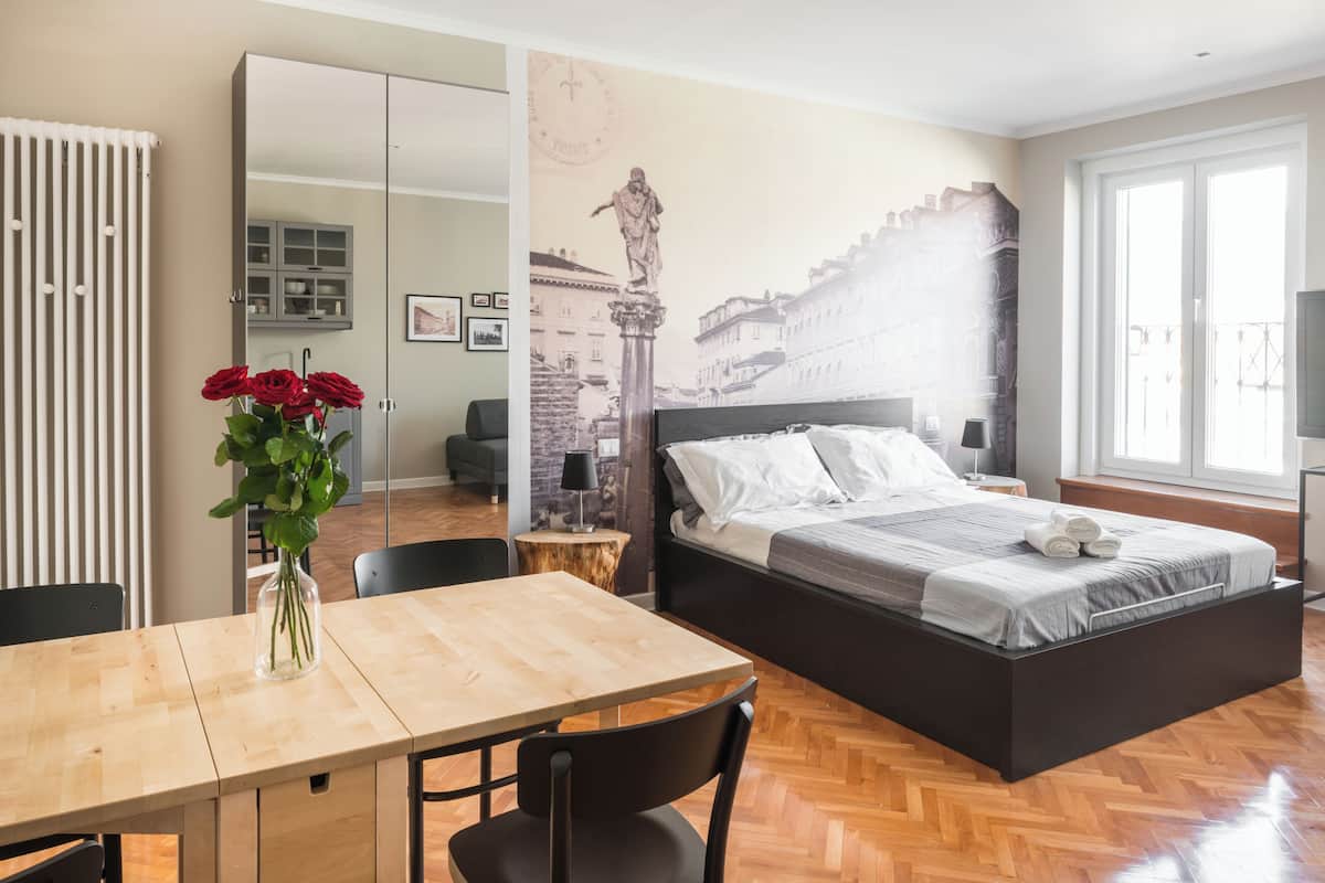 Quarto com cama, mesa de madeira, cadeiras, um vaso com flores e um mural na parede.