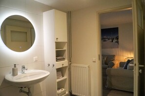 A bathroom with a sink, mirror, and storage cabinets. There is a radiator and a doorway leading to a living area with a couch and a lamp.
