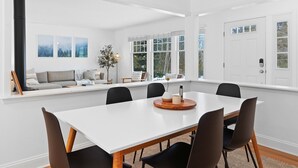 Un coin repas avec une table blanche, des chaises noires et un banc près de la fenêtre, d’où l’on peut admirer les arbres.