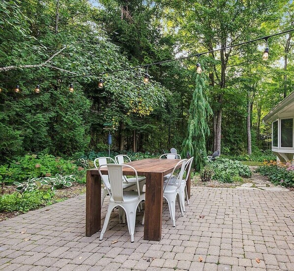 An outdoor dining area with a brick patio, a wooden table, and white chairs.