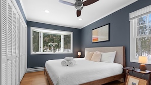 Une chambre avec un grand lit, un ventilateur de plafond, une fenêtre donnant sur les arbres et une table de chevet avec une lampe.