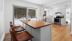 Une cuisine moderne dotée d’un îlot central, de meubles blancs et d’une grande fenêtre offrant une vue sur les arbres.