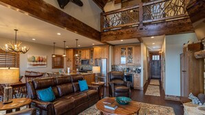 A spacious living area with a leather sofa, a coffee table, and a dining area with wooden chairs. A kitchen with wooden cabinets and a stone backsplash is visible in the background. - Whitetail Ridge Lodge - Eagles Nest, long range views, three masters (Banner Elk)