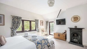 A bedroom with a bed, a dresser, a fireplace, a television, and a window with curtains. - The Willows - Woodland Cottage with Log Burner (Christchurch)