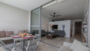 Un vaste espace de vie comprenant une table à manger pour deux personnes, un canapé et une table basse. Une porte-fenêtre coulissante donne accès à un balcon.