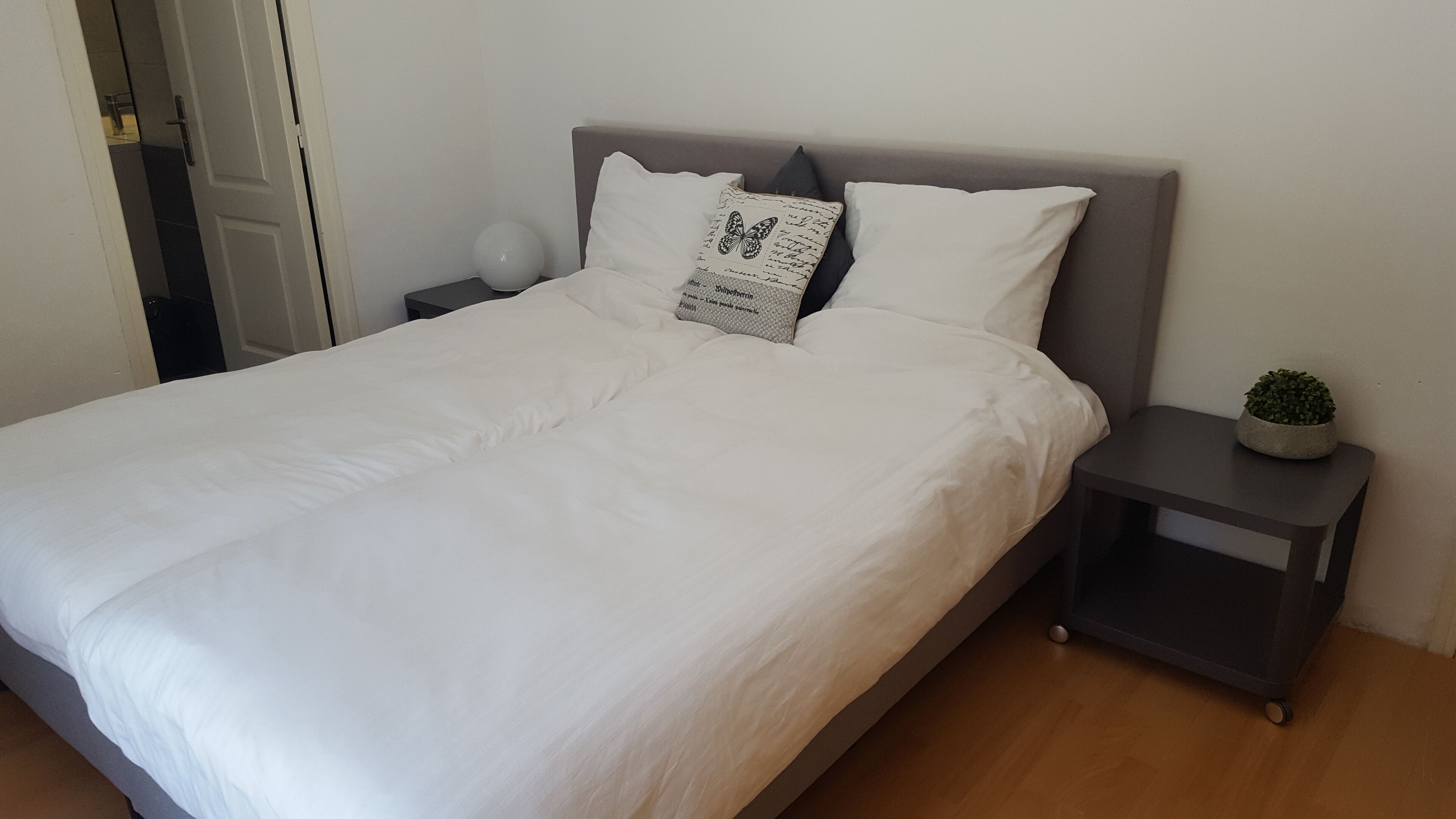 A neatly made bed with white linens, a grey headboard, and two bedside tables with a small plant on one.