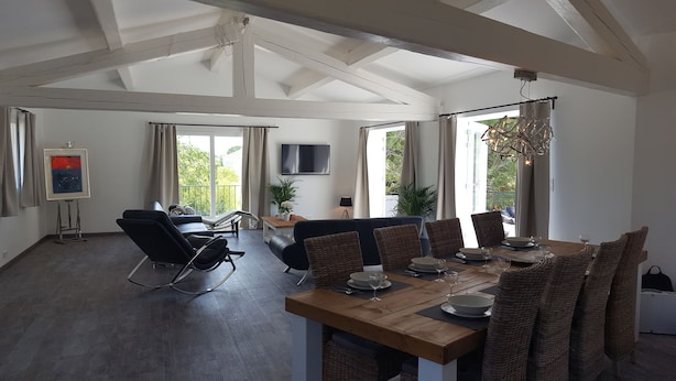 Amplia sala de estar con una mesa de comedor para cuatro personas, un sillón reclinable, un televisor de pantalla plana montado en la pared y un balcón con cortinas.