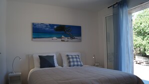 Un dormitorio con cama, mesita de noche con lámpara, cuadro de una playa y vista a zonas verdes por la ventana.