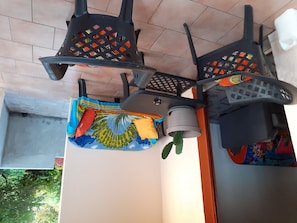 Una habitación con piso de baldosas, ventilador de techo y estante en la pared con un textil colorido y un felpudo enrollado.