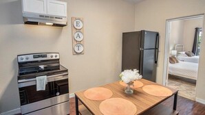 Eine kleine Küche mit Herd, Backofen und Kühlschrank, ein Holztisch mit Platzsets und ein Bett, das durch eine geöffnete Tür sichtbar ist.